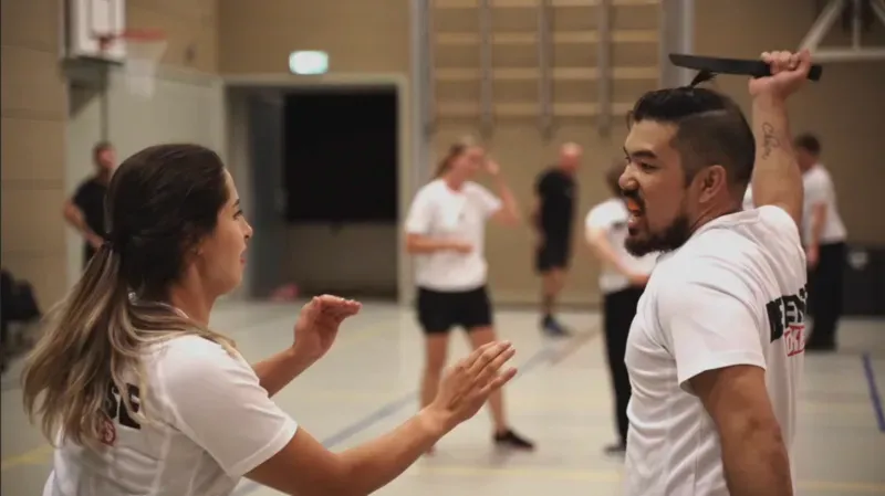 Mesverdediging tijdens Krav Maga-training: een deelneemster houdt de afstand terwijl de aanvaller met een trainingsmes uithaalt